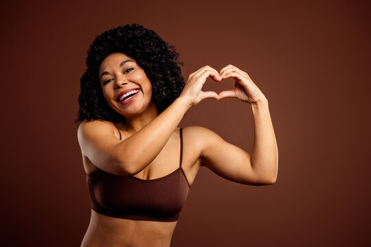 Joyful woman with curly hair forming a heart shape with hands on a brown background embodying positivity and confidence