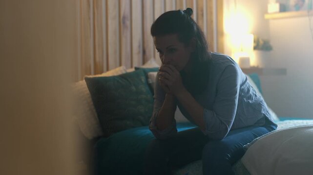 Woman sitting on edge of bed with hands clasped near mouth, anxiety and deep reflection, symbol of emotional struggle and mental fatigue in private home setting