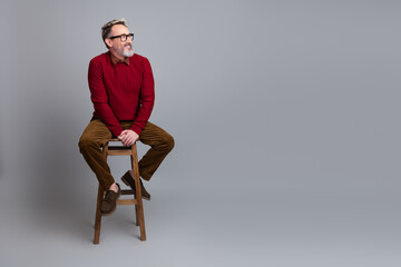 Confident middle-aged businessman sitting stylishly on a wooden stool against a gray background...