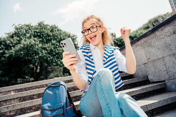 Happy student enjoying outdoor leisure with her phone on a sunny day in the city while sitting on...