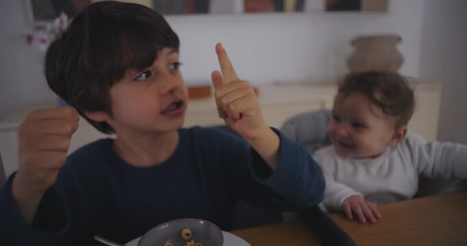 Young boy eating breakfast cereal while counting with his fingers, sitting beside his baby sibling at the table, showing learning, curiosity, and warm family interaction