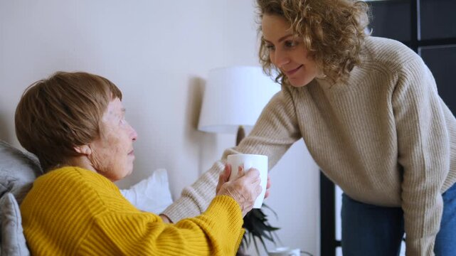 Compassionate young daughter taking care of her sick elderly mother in bed, giving her a warm cup of tea and talking, showing love, support, and family care during illness