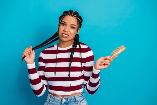 Young woman with braided hair wearing striped top stands against blue background holding a hairbrush and showing a casual stylish lifestyle portrait - Powered by Adobe