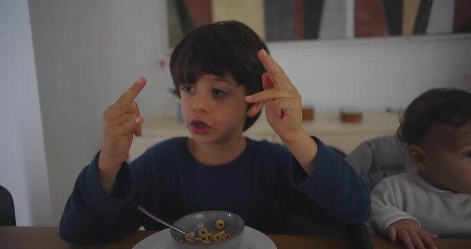 Young boy sitting at the table counting with his fingers while his baby sibling watches and smiles, expressing curiosity, learning, and the joy of shared family moments