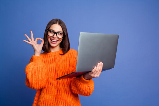 Young woman in an orange sweater gesturing okay while holding a laptop, expressing excitement against a blue background