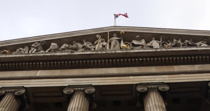 Low angle view of a grand neoclassical facade in London, with the Union Jack flying above carved stone figures and columns. The scene suggests empire, cultural authority and national identity.