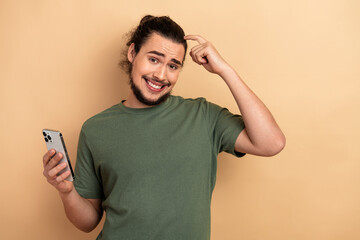 Young man with smartphone smiles and points to head in casual green t shirt against beige background