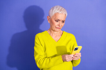Older woman in bright yellow sweater looking at her phone with a thoughtful expression against a...