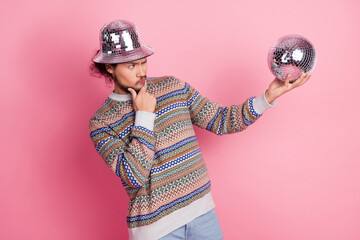 Trendy young man in a fashionable sweater holding a mirror disco ball against a pastel pink...