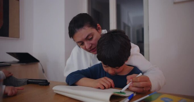 Mother helping son draw in notebook at table showing caring family bond parenting guidance and authentic everyday learning moment at home indoors