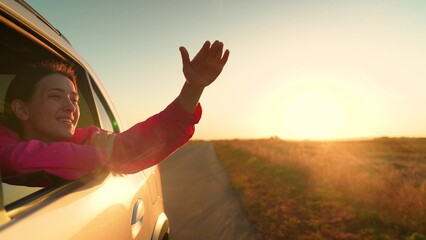 Young female passenger catches wind from car window. Beautiful female student drives car along road, sunset. Family trip, teenage girl travel by car. Family vacation. People traveling. Woman freedom