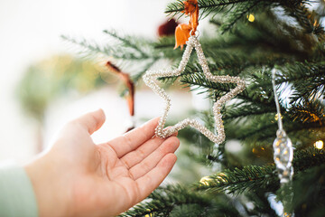 Hand holding stylish vintage star on Christmas tree branch close up. Woman hanging modern ornament, decorating xmas tree with lights. Winter holiday banner, atmospheric christmas time
