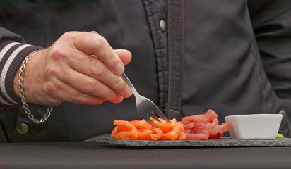 A person sampling slices of fresh salmon and tuna served on a black slate plate with a small bowl of sauce at a farmers market in Prague.