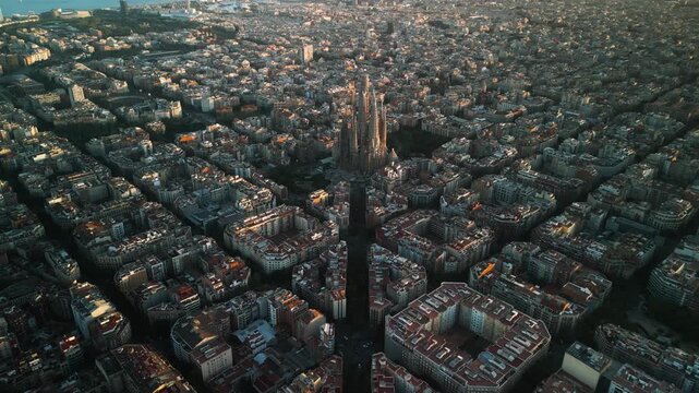 4K drone aerial view of Sagrada Familia in Barcelona at sunset, showing golden light over Eixample district and iconic grid city pattern, Catalonia, Spain