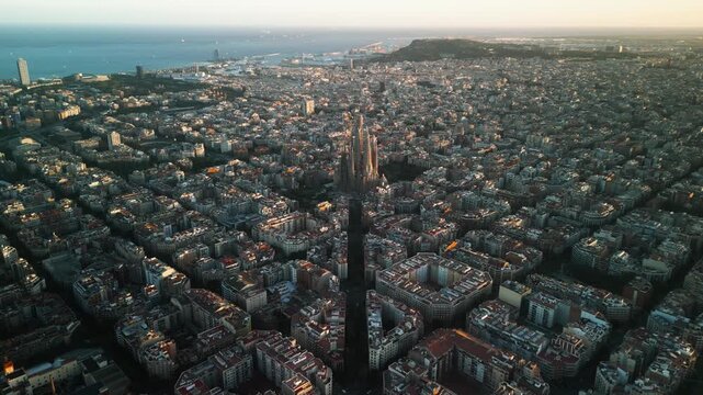 4K drone aerial view of Sagrada Familia in Barcelona at sunset, showing golden light over Eixample district and iconic grid city pattern, Catalonia, Spain