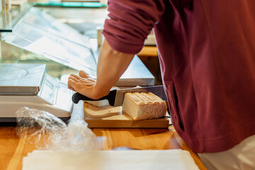 Delicatessen shop interior: hands slicing artisan goat cheese with slicer. Small business typical local products. Counter with cheeses. Welcoming authentic traditional commerce atmosphere.