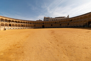 An empty bullring with a large sandy arena floor is surrounded by arched seating under a clear blue sky. © DBA