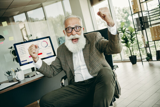 Senior businessman celebrates success in stylish office environment with a computer and financial data on screen