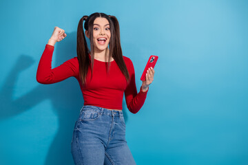 Excited young woman in casual outfit holding smartphone celebrates success against vibrant blue...