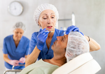 Adult man applying rejuvenating treatments by mature female doctor in cosmetology office. Nurse...
