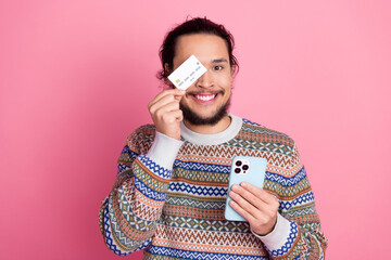 Happy young man holding smartphone and credit card, wearing a multicolored sweater, with a cheerful expression against a pink background.