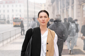 Businesswoman in black coat standing on sidewalk in city. Woman has confident expression with focused gaze. Soft light highlights her face against blurred background of buildings 