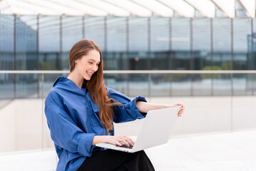 Smiling businesswoman in blue shirt working on laptop in front of modern glass office building. Enjoying her work in bright environment