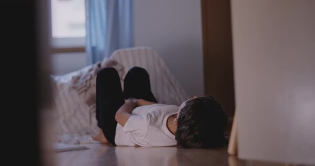 Child curled up on floor beside bed with arms resting on chest, looking tired and bored, expressing disconnection, restlessness, and lack of interest in quiet moment
