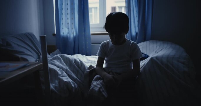 Boy sitting on bed with head bowed in dim bedroom, looking down in sadness and reflection, symbolizing loneliness, despair, emotional struggle, and childhood isolation