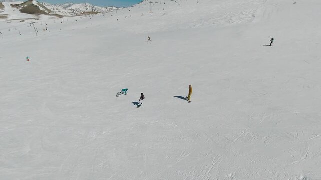 Tracking aerial distant view of skiers and snowboarders on a wide open mountain slope. One instructor helps a beginner balance on snow while others ride alone, small against white winter  landscape.