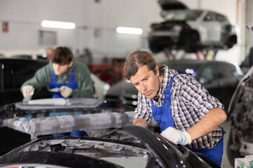 Focused experienced middle aged mechanic inspecting detached car bumper in workshop, scrutinizing damage, contemplating necessary steps for restoration or repair..