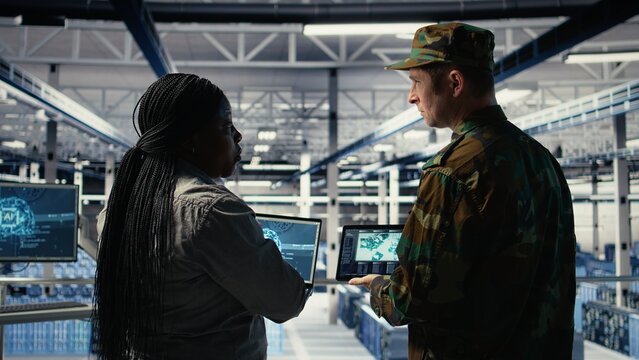 Military commander and female programmer in data center tracking coordinates using notebook software. Soldier and african american woman in server hub analyzing reconnaissance data, camera A