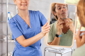 Adult man examining his face after cosmetology procedures in mirror reflection in aesthetic medicine office..