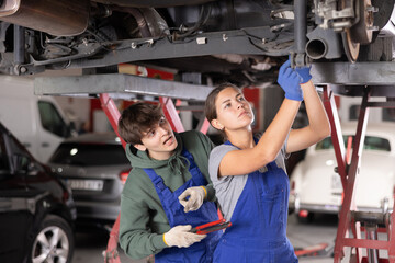 Young female auto mechanic in blue overalls standing with assistant under car on lift in service workshop, confidently repairing undercarriage