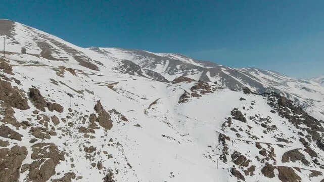 Aerial tracking shot across steep winter mountains. Jagged rocks break through the melting snow, revealing brown earth and winding paths. Remote altitude, harsh beauty, exposure, silence, endurance.