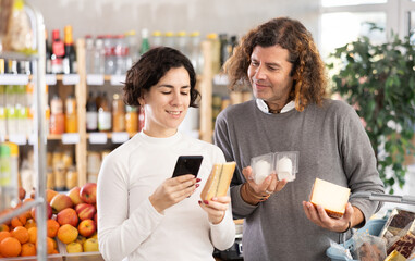 Spouses gets acquainted with drink scan QR code, take reading cheese using phone camera in shop. Client view varieties of cheese, inspect manufacturer information using mobile app