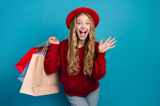 Adorable young woman wearing a red sweater holding shopping bags while smiling joyfully against a bright blue background