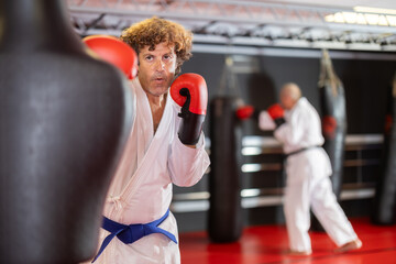 Middle-aged karate student , blue belt holder, ın red boxıng gloves delivering controlled punch to mannequin-style bag, refining technique and timing in class during casual routine..