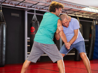 Elderly man and adult man practice self-defense techniques at gym