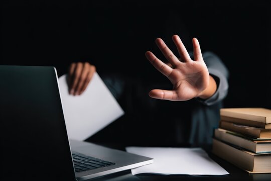 Person showing stop gesture, refusing to accept a document or offer at a desk with a laptop and books