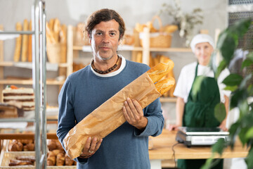 Man visitor with baguettes in paper bag eko-package, stands near showcase in bakery. Client bought...