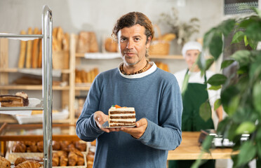 Man customer holds disposable plate with cake in sales area of bakery, takes out fresh pastries...
