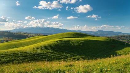 Fototapeta premium Lush green rolling hills under a bright blue sky with fluffy white clouds and distant mountains