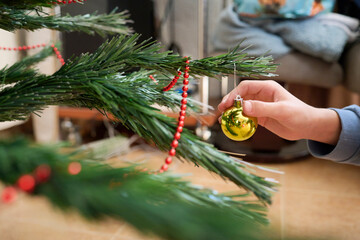Hand of an unrecognizable child places a Christmas ball on a branch of the plastic tree