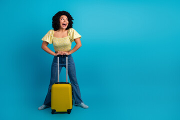 Happy young woman with black curly hair holds yellow suitcase on blue background, excited to travel and explore