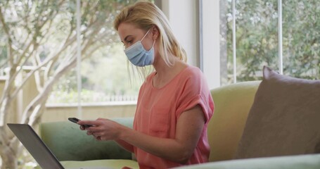 Holding smartphone, woman wearing surgical mask on couch by window with open laptop, copy space