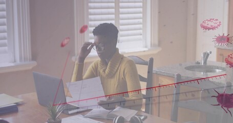 Woman in mustard sweater with glasses reviewing printouts at kitchen table, with laptop headphones