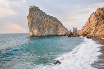 High coastal rock near the sea, waves on the shore, seascape with turquoise water, sunset light, natural beauty of the coastline