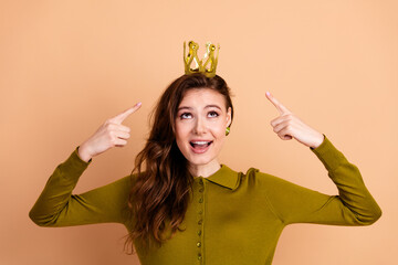 Young woman wearing a crown points to her playful look in a casual khaki sweater against a peach...