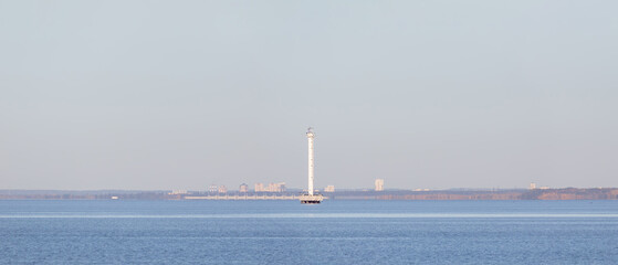 A tall white lighthouse rises from the calm blue sea near the coastline under a clear sky, with...
